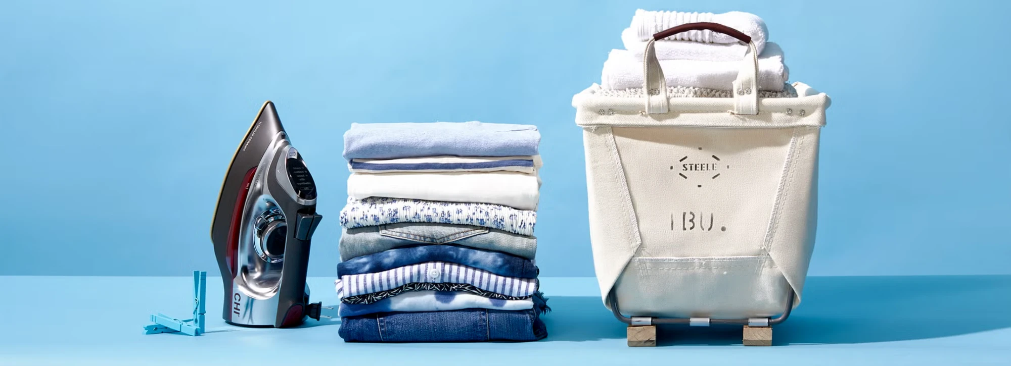 A neat stack of freshly washed and folded shirts and jeans between an iron and a canvas laundry bin